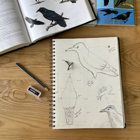 Notebook with bird illustrations on a wooden surface, next to an open book and a pencil.